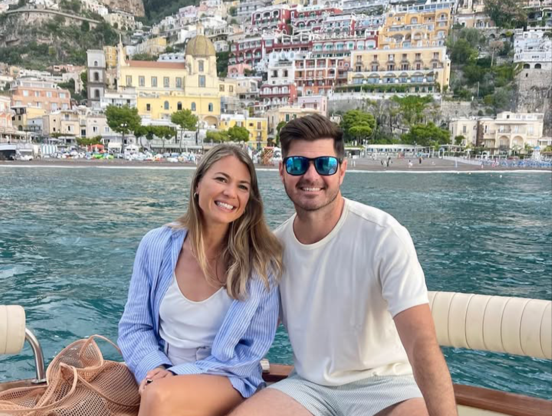 A smiling woman and man sitting on a boat, with a coastal town and hillside buildings in the background.