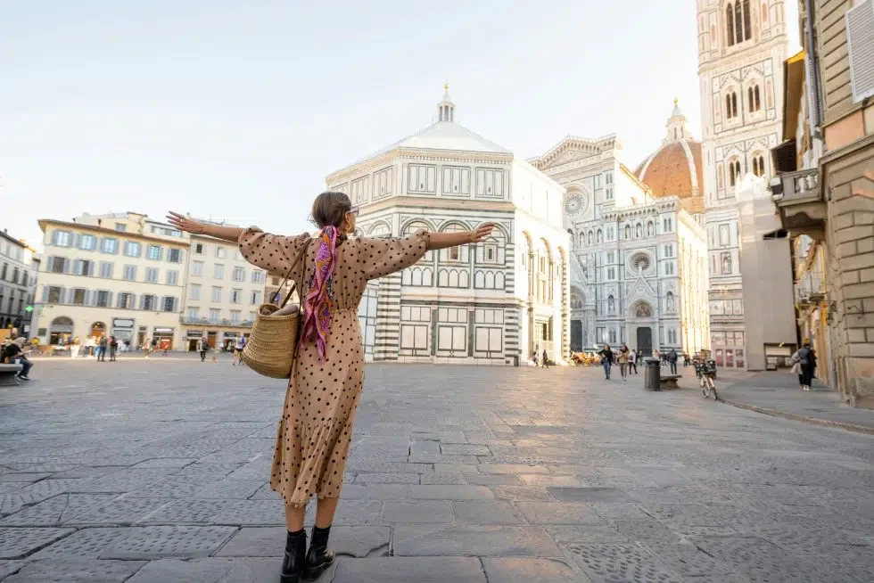 Woman standing with arms outstretched in Piazza del Duomo in Florence, facing the Baptistery and Florence Cathedral, wearing a polka dot dress and scarf on a bright, open morning.