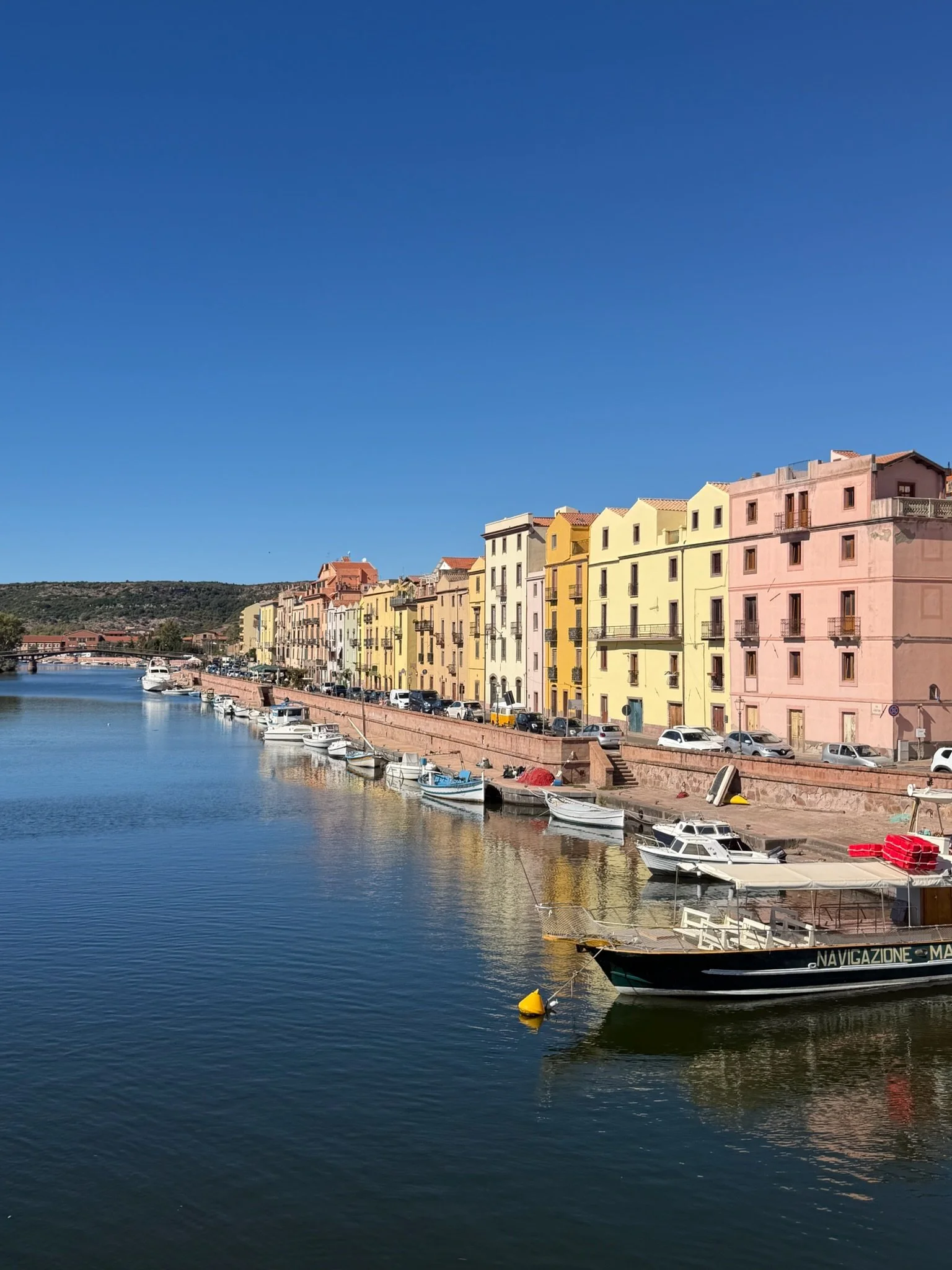 Colorful pastel buildings lining the Temo River in Bosa, Sardinia, Italy, with fishing boats moored along the waterfront beneath a clear blue sky.