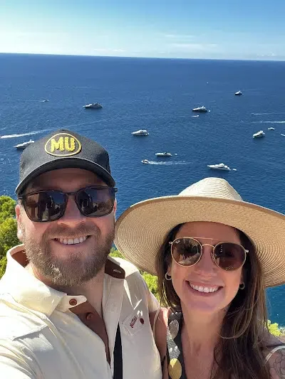 A smiling couple taking a selfie on a sunny day near the ocean, with multiple boats visible on the water behind them. The man is wearing sunglasses, a cap, and a light-colored shirt, and the woman is wearing sunglasses and a wide-brimmed hat.