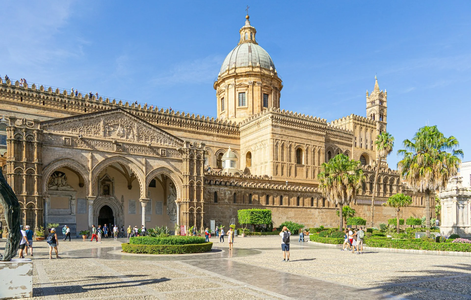 Palermo Cathedral in Palermo, Sicily, featuring its grand Norman-Arab architecture, soaring dome, Gothic details, palm trees, and visitors exploring the sunlit piazza beneath a bright blue sky.