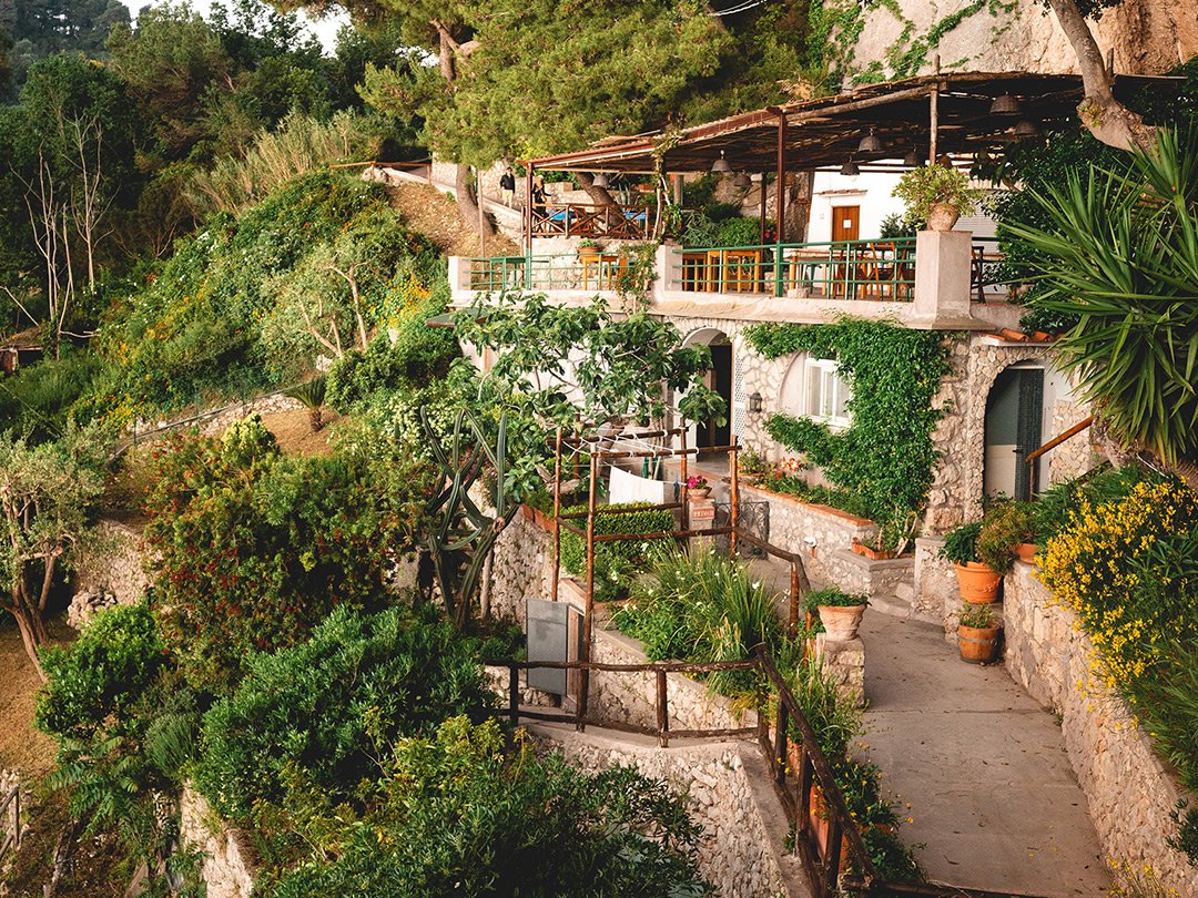 A multi-level stone and wood house built into a lush hillside, with terraces and balconies decorated with potted plants and greenery.