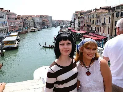 Two women standing on a bridge over a canal in Venice, Italy, with colorful buildings lining the water and boats in the background.