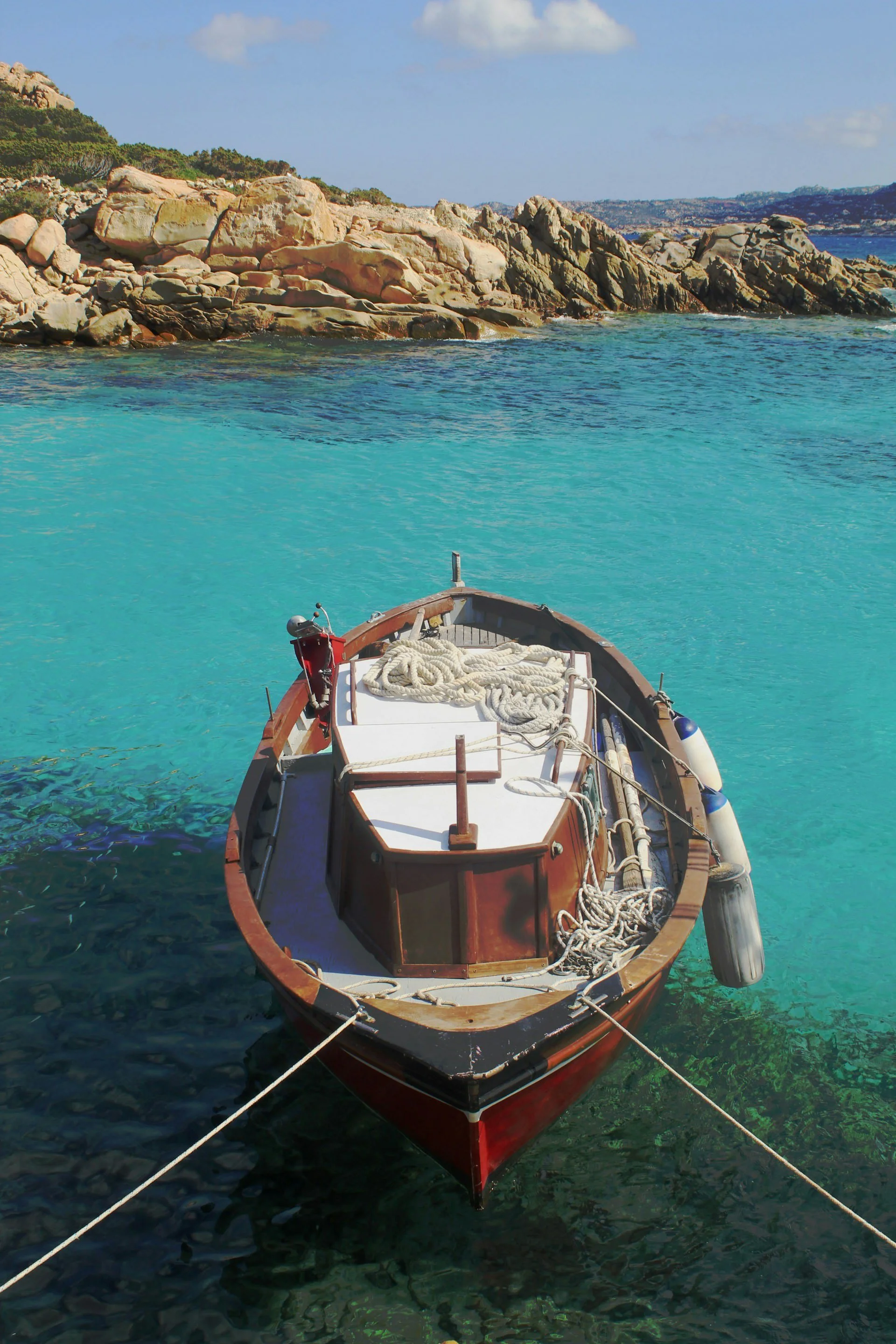 Small wooden boat moored in clear turquoise water along a rocky Sardinian coastline, with sunlit stone formations and calm Mediterranean sea.