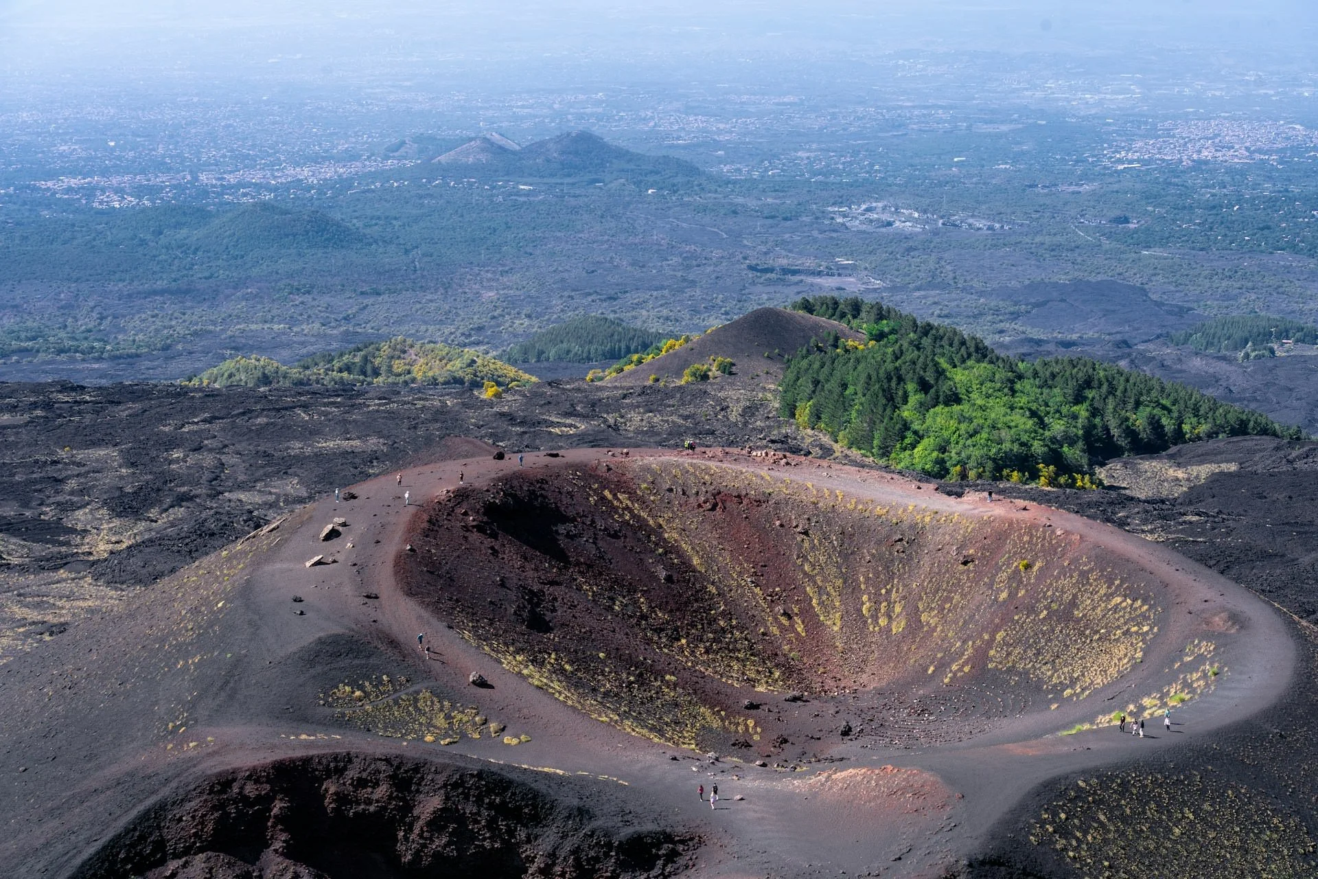 Aerial view of Mount Etna’s volcanic crater with dark lava fields, green forest patches, and hikers walking along the rim in Sicily.