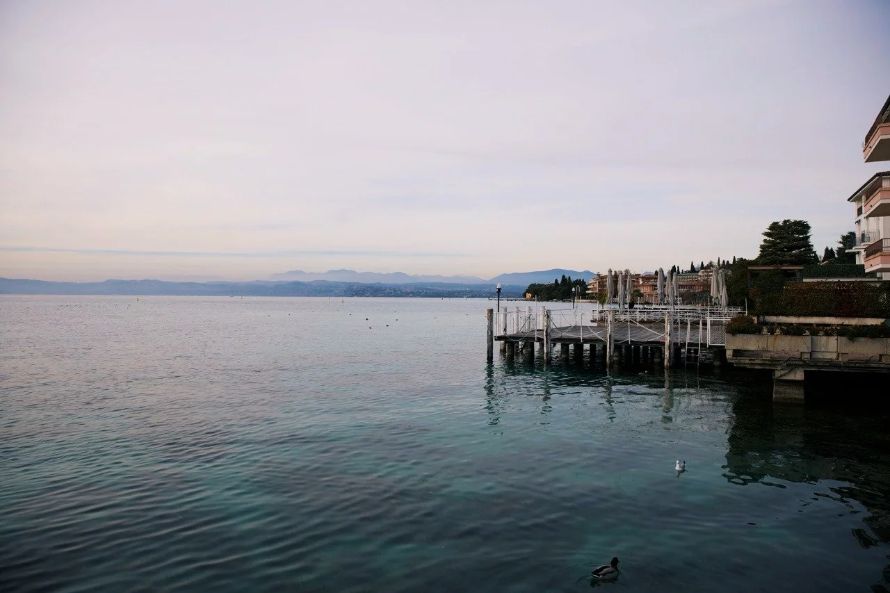 Peaceful waterfront view of Lake Garda in Sirmione, Italy, with calm blue water, lakeside docks, and distant mountains under a soft pastel evening sky.