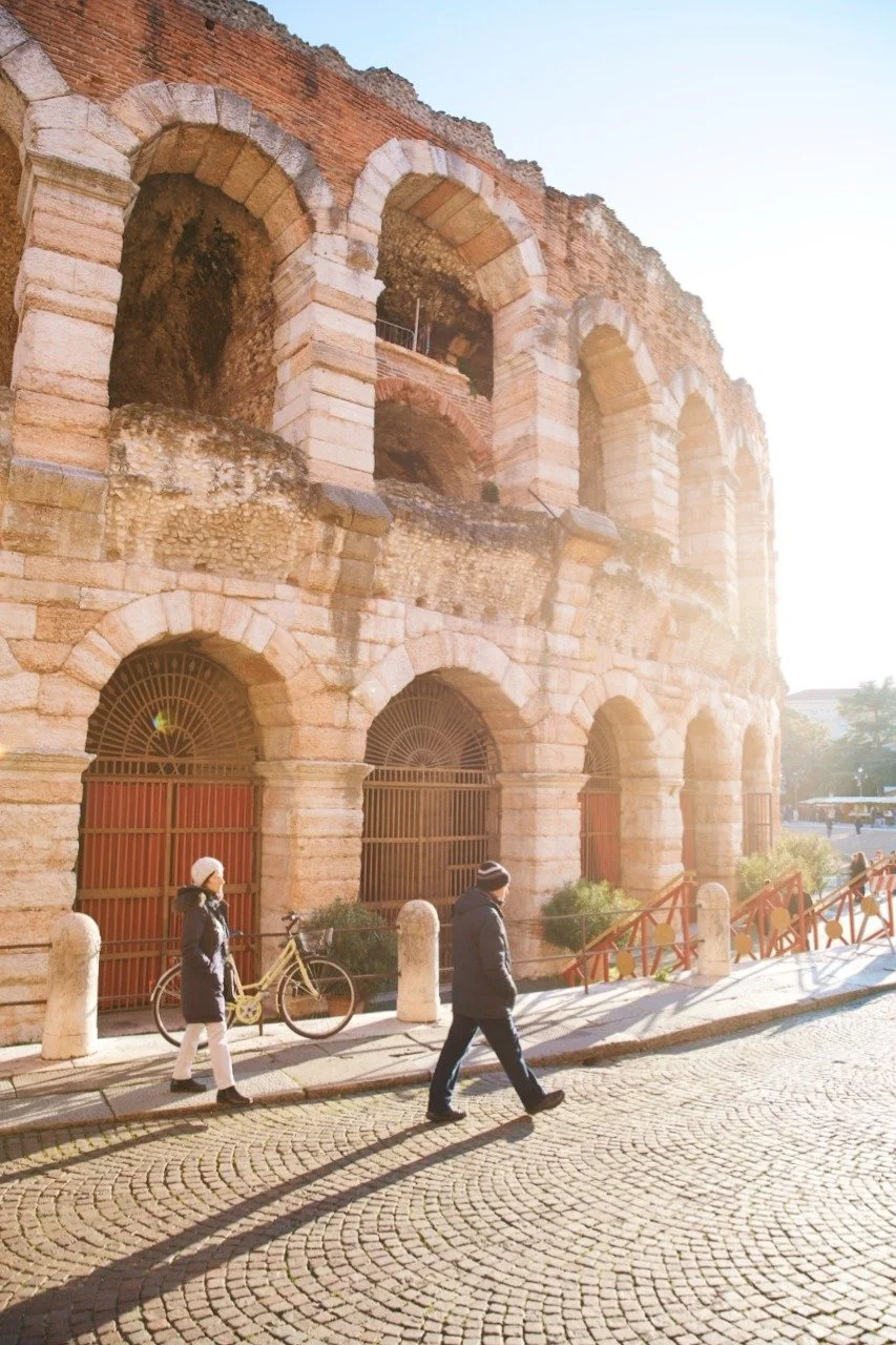 Sunlight streaming across the ancient Arena di Verona amphitheater in Verona, Italy, with pedestrians walking along the cobblestone street beside the historic arches.
