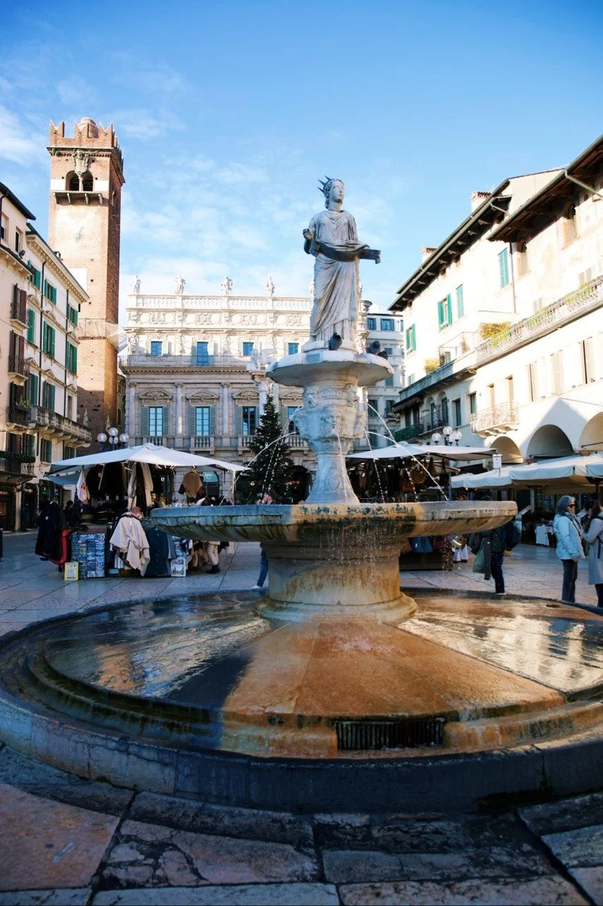 Fountain in Piazza delle Erbe in Verona, Italy, surrounded by historic buildings, market stalls, and winter sunlight under a clear blue sky.