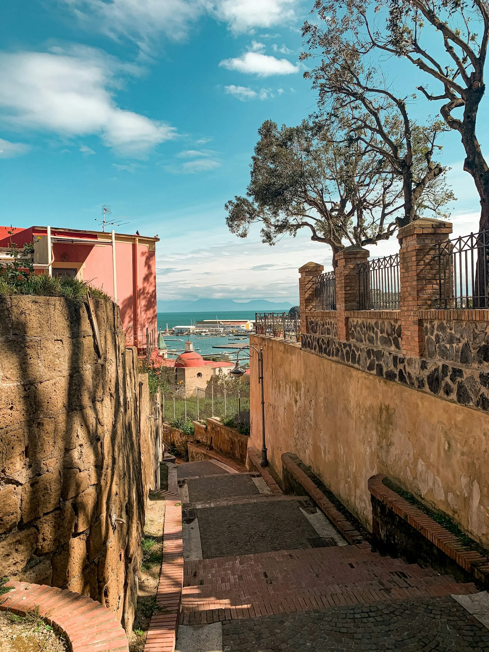 Stone pathway descending between historic buildings toward the waterfront with views of the Bay of Naples under a blue sky.
