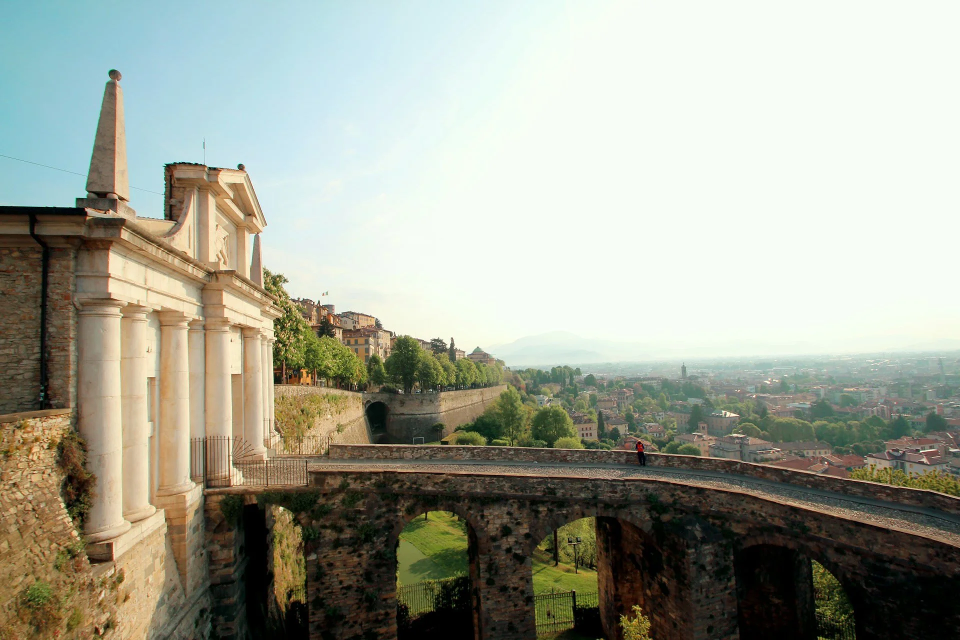 Panoramic view of Bergamo, Italy featuring historic stone arches, Renaissance architecture, and the city skyline glowing in soft morning light.
