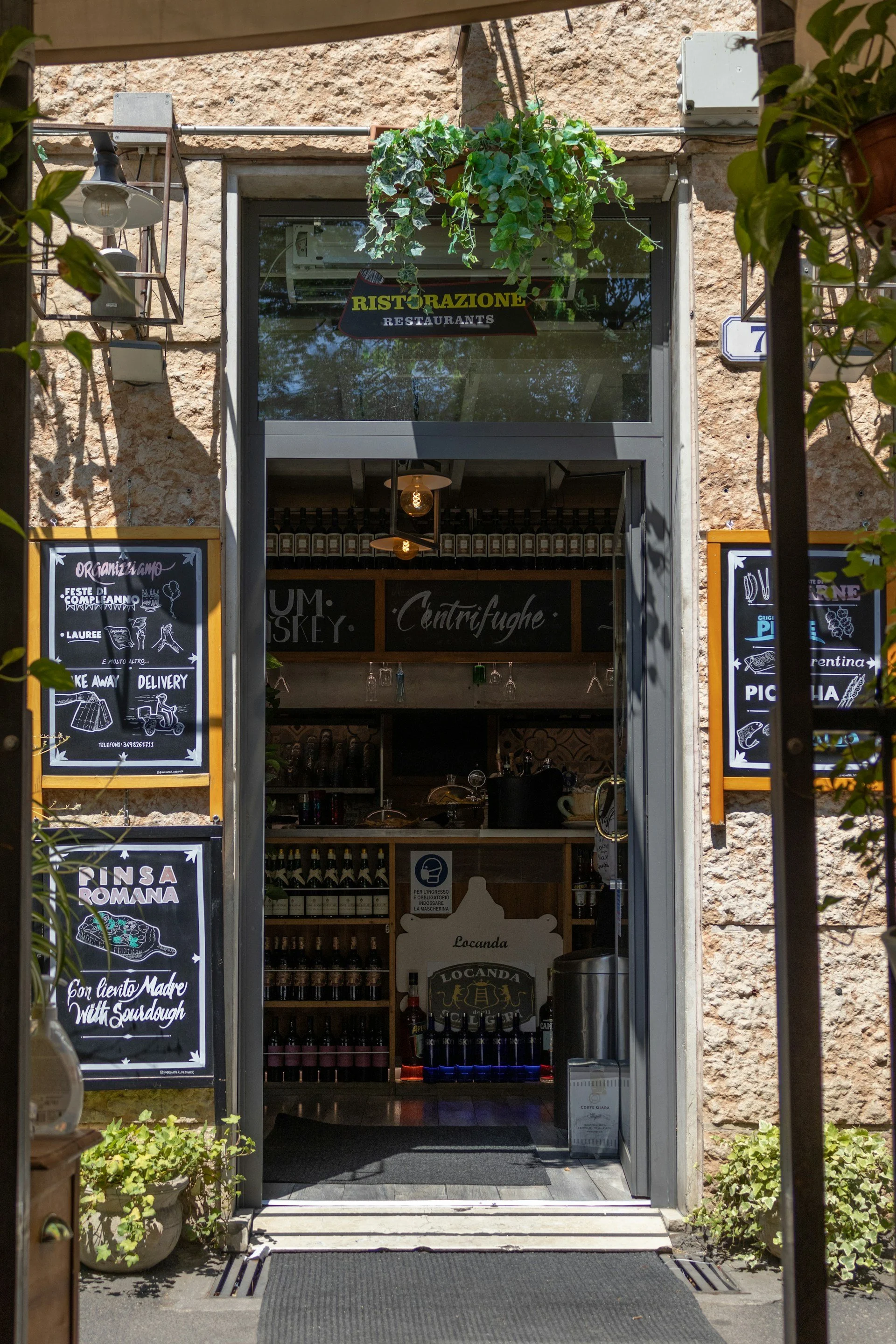 Entrance to a charming Italian restaurant in Verona featuring rustic stone walls, hanging greenery, wine displays, and traditional menu boards advertising local specialties and pizza.