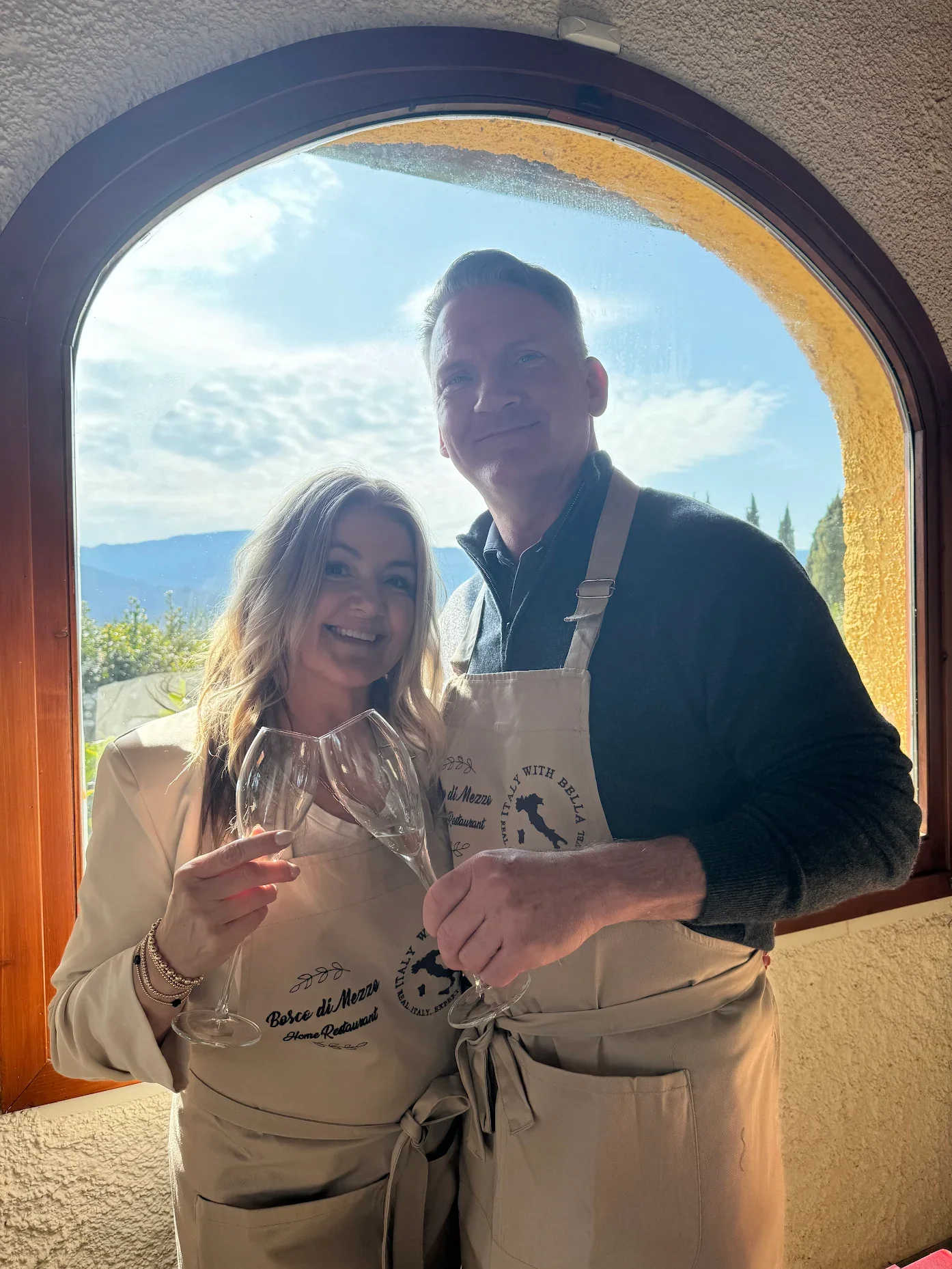 A man and woman inside a restaurant, standing in front of a window with a scenic outdoor view. They are dressed in aprons, holding wine glasses, and smiling at the camera.
