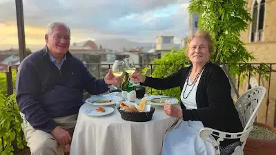An elderly couple sitting at a table outdoors, enjoying drinks and smiling.