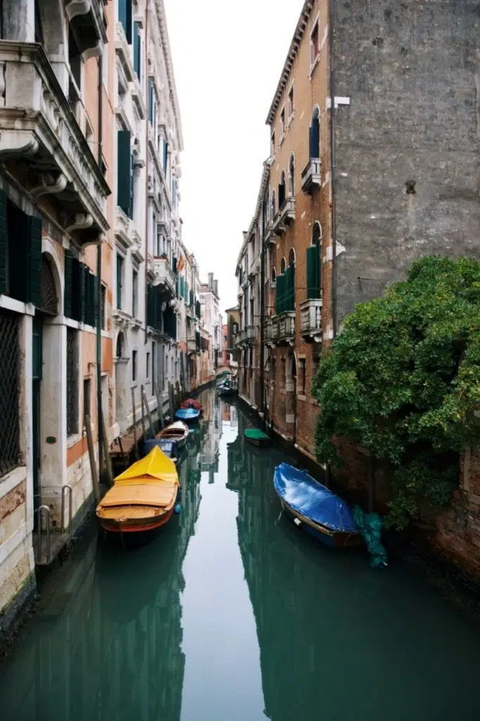 Narrow Venice canal lined with historic buildings, featuring small boats moored along calm green water, with colorful boat covers and a quiet, atmospheric perspective down the canal.
