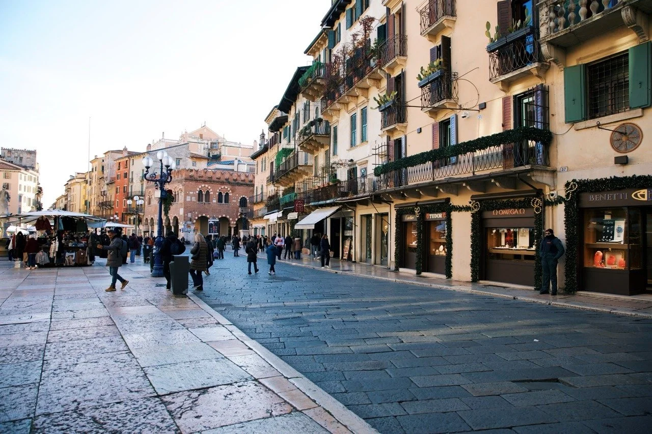 Shoppers and locals strolling through Piazza delle Erbe in Verona, Italy, lined with elegant historic buildings, balconies, and boutique storefronts on a bright winter afternoon.