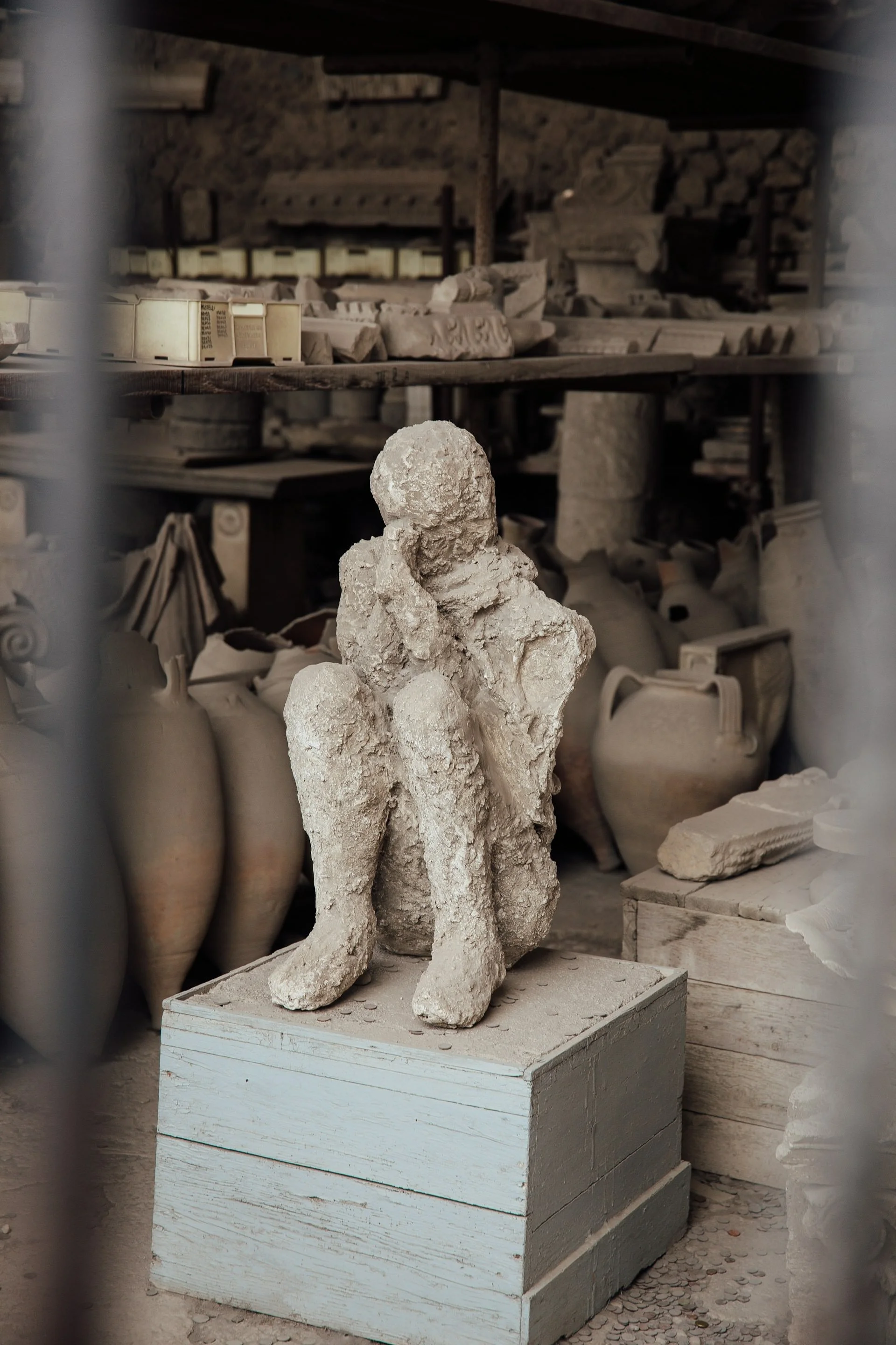 Plaster cast of a Pompeii victim seated with knees drawn in, displayed among ancient pottery and artifacts inside an archaeological storage room