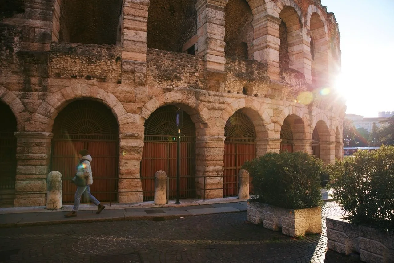 Sunlight streaming across the ancient stone arches of the Arena di Verona as a traveler walks along the quiet street in Verona, Italy.