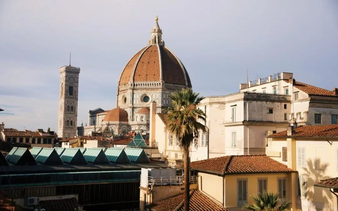 View of the Florence skyline featuring the Duomo with its terracotta dome, Giotto’s Campanile, palm trees, and sunlit rooftops under a clear sky.