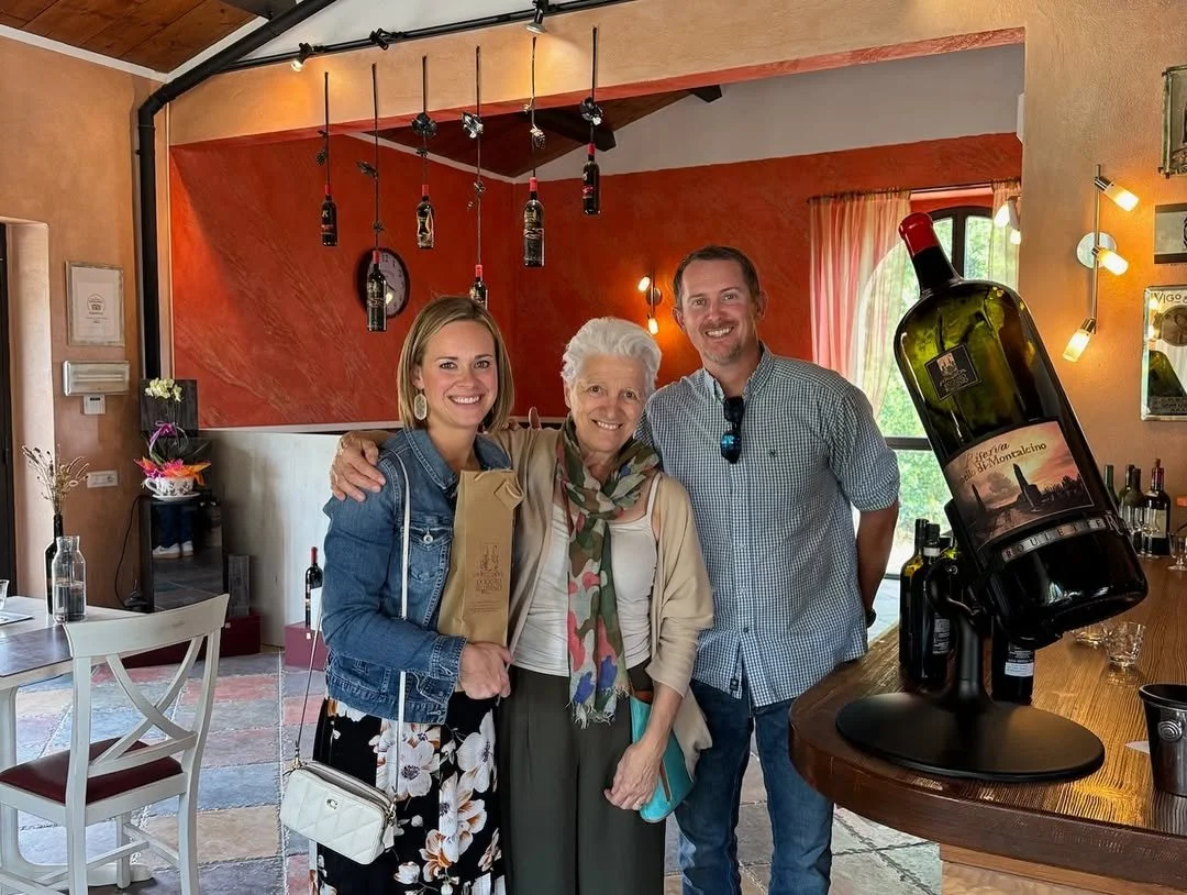 Three people, two women and one man, smiling and posing for a photo inside a wine bar or restaurant. One woman is holding a paper bag, and there is a large wine bottle display on the bar to their right.