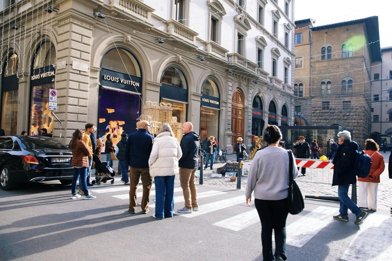 Pedestrians crossing a busy street in Florence, Italy, near luxury storefronts and historic architecture during the holiday season, with street musicians performing in the background.