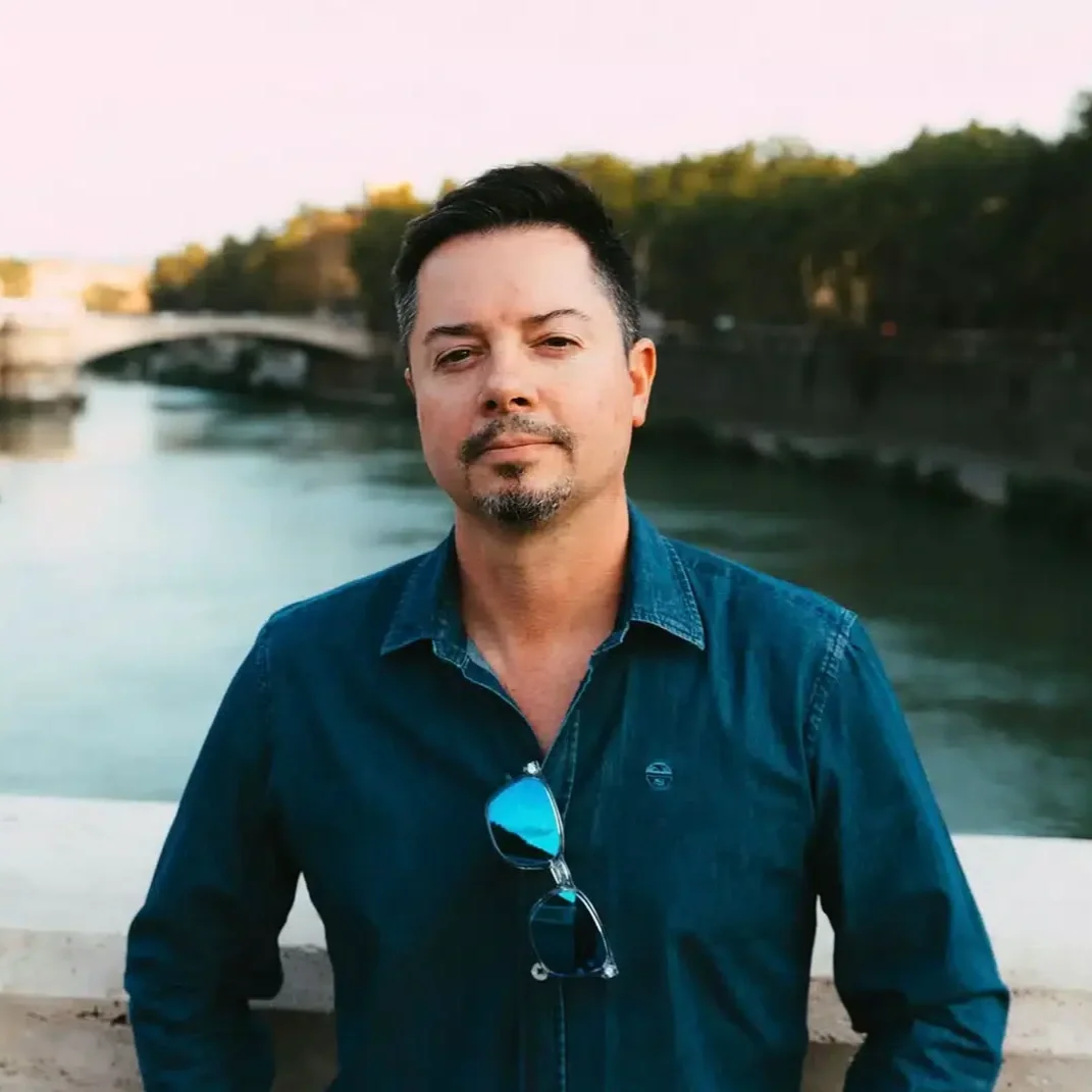 A man with dark hair, a trimmed beard, wearing a blue denim shirt with sunglasses hanging from the collar, standing in front of a river with a bridge in the background during sunset.