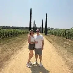A couple standing on a dirt road with tall cypress trees in the background, surrounded by a field of crops.