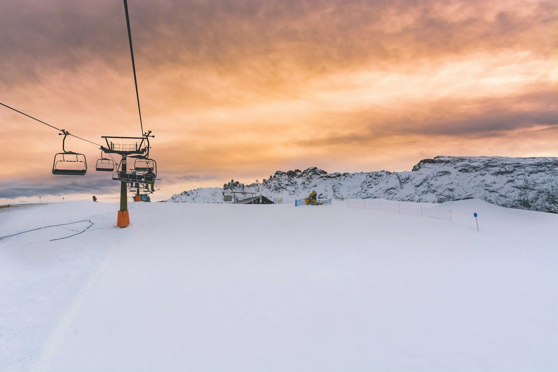 Snow-covered ski slope at sunset with empty chairlifts, rugged mountain peaks, and a glowing pastel sky in the background.