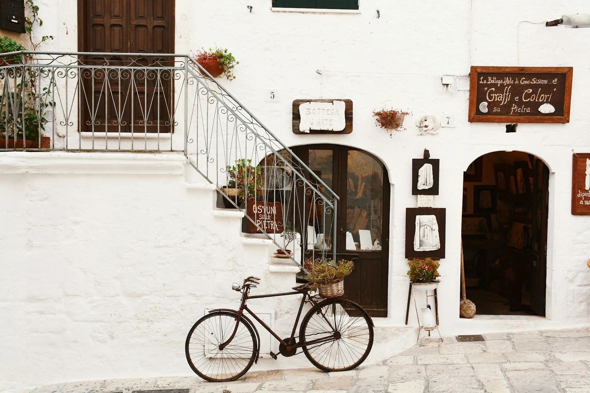 Whitewashed building with iron staircase, potted plants, and a vintage bicycle parked outside a small artisan shop in a charming Italian village street.