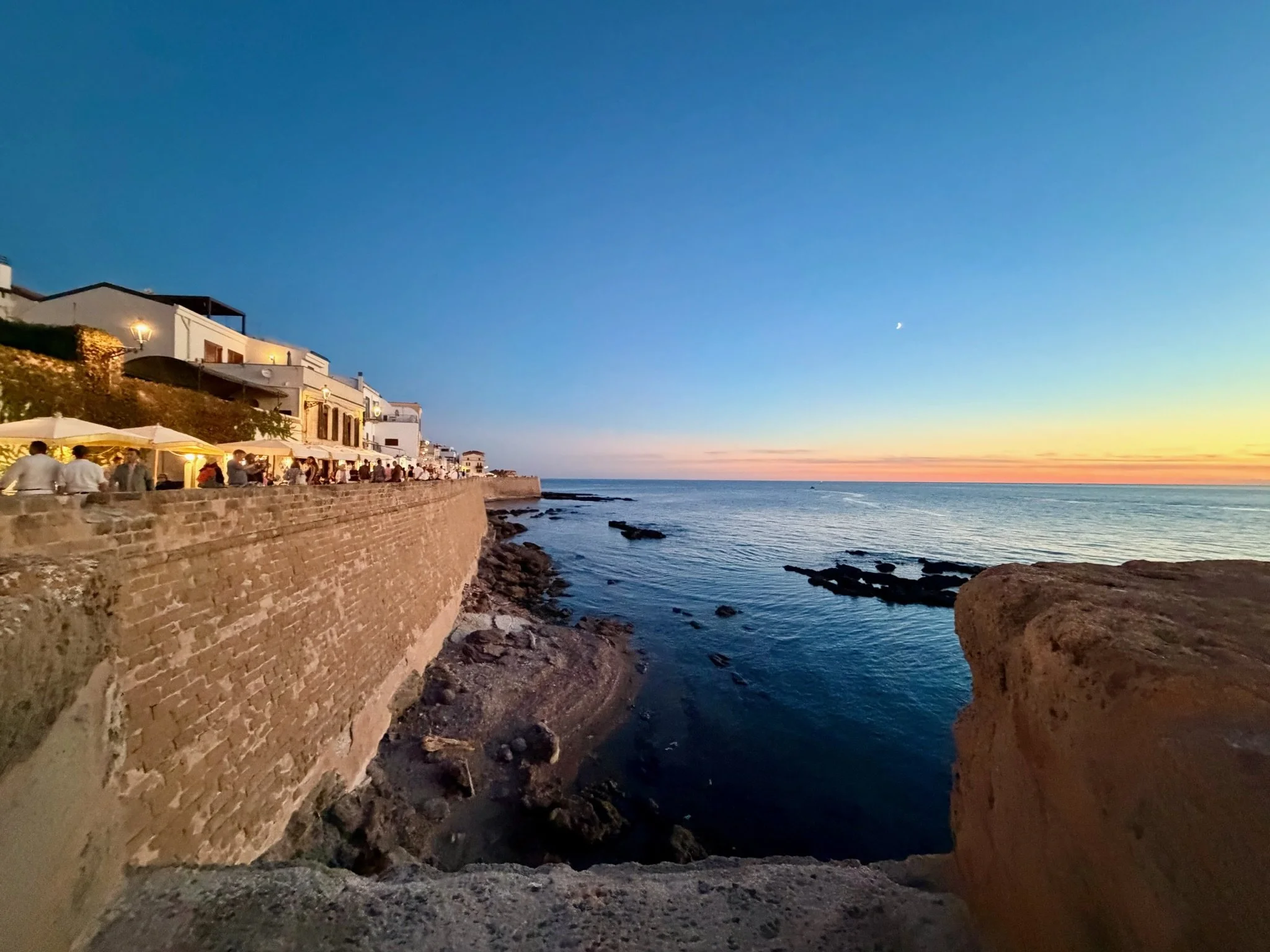 Sunset over the Mediterranean Sea in Alghero, Sardinia, Italy, with waterfront restaurants, historic stone walls, and a crescent moon in the evening sky.