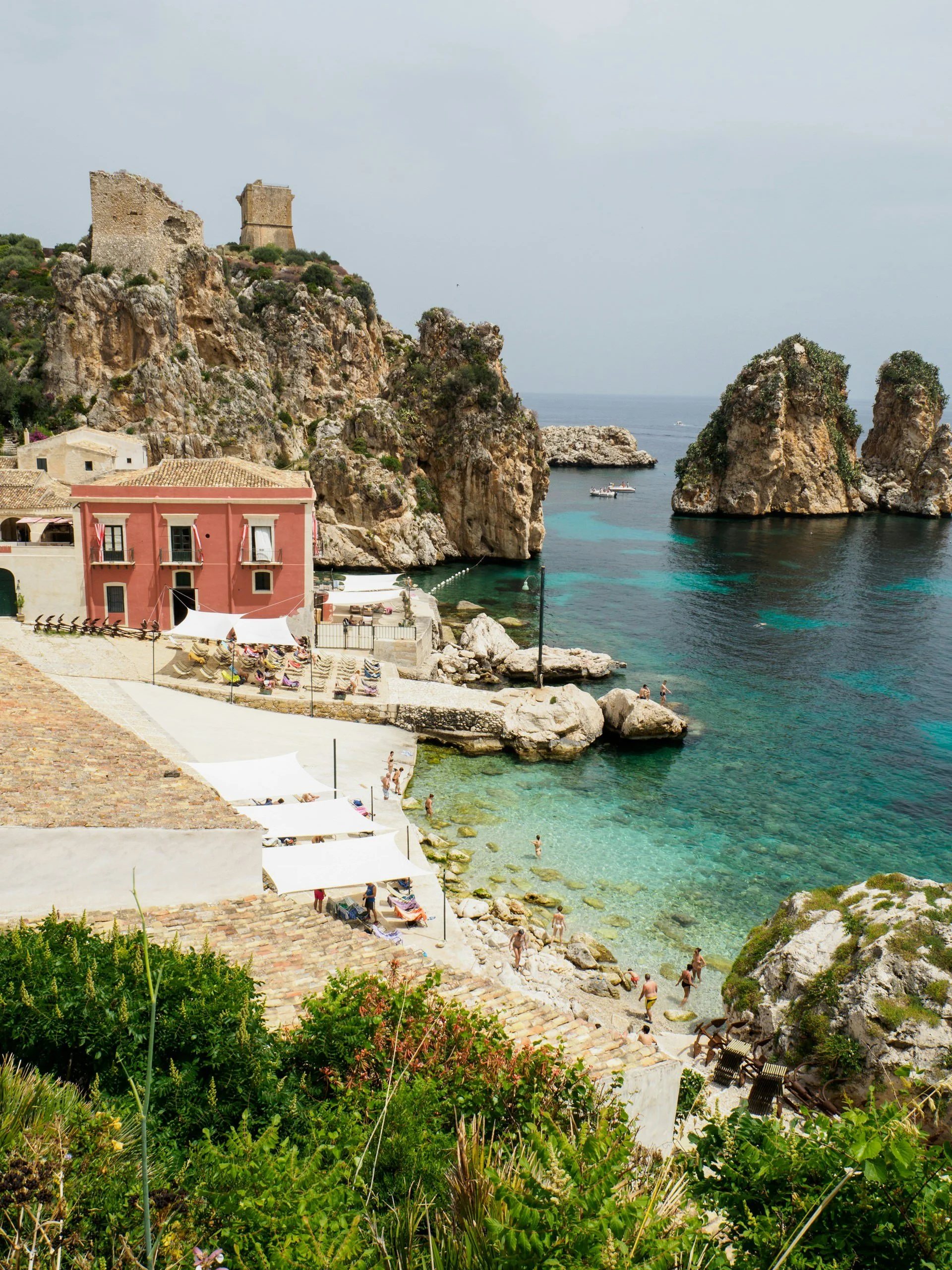 Turquoise waters and dramatic sea stacks at Tonnara di Scopello in Sicily, with sunbathers along a rocky cove and a historic seaside building overlooking the Mediterranean.