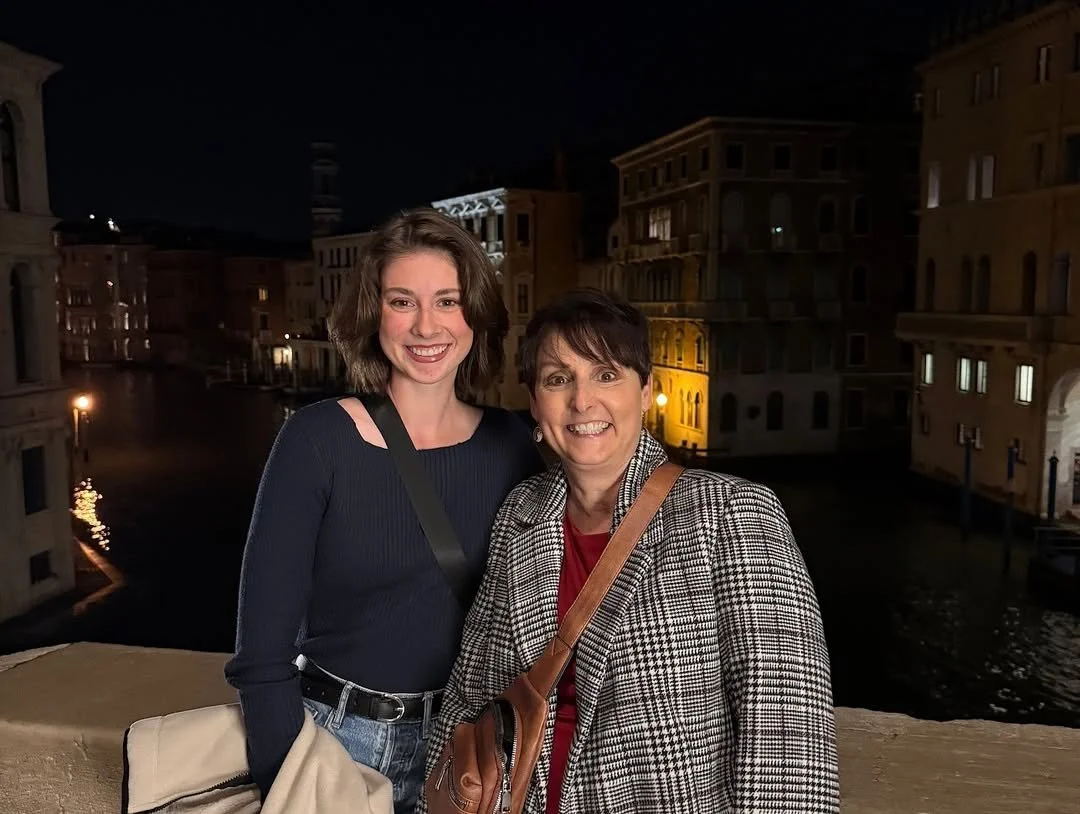 Two women smiling on a bridge in Venice at night with illuminated historic buildings and canals in the background.