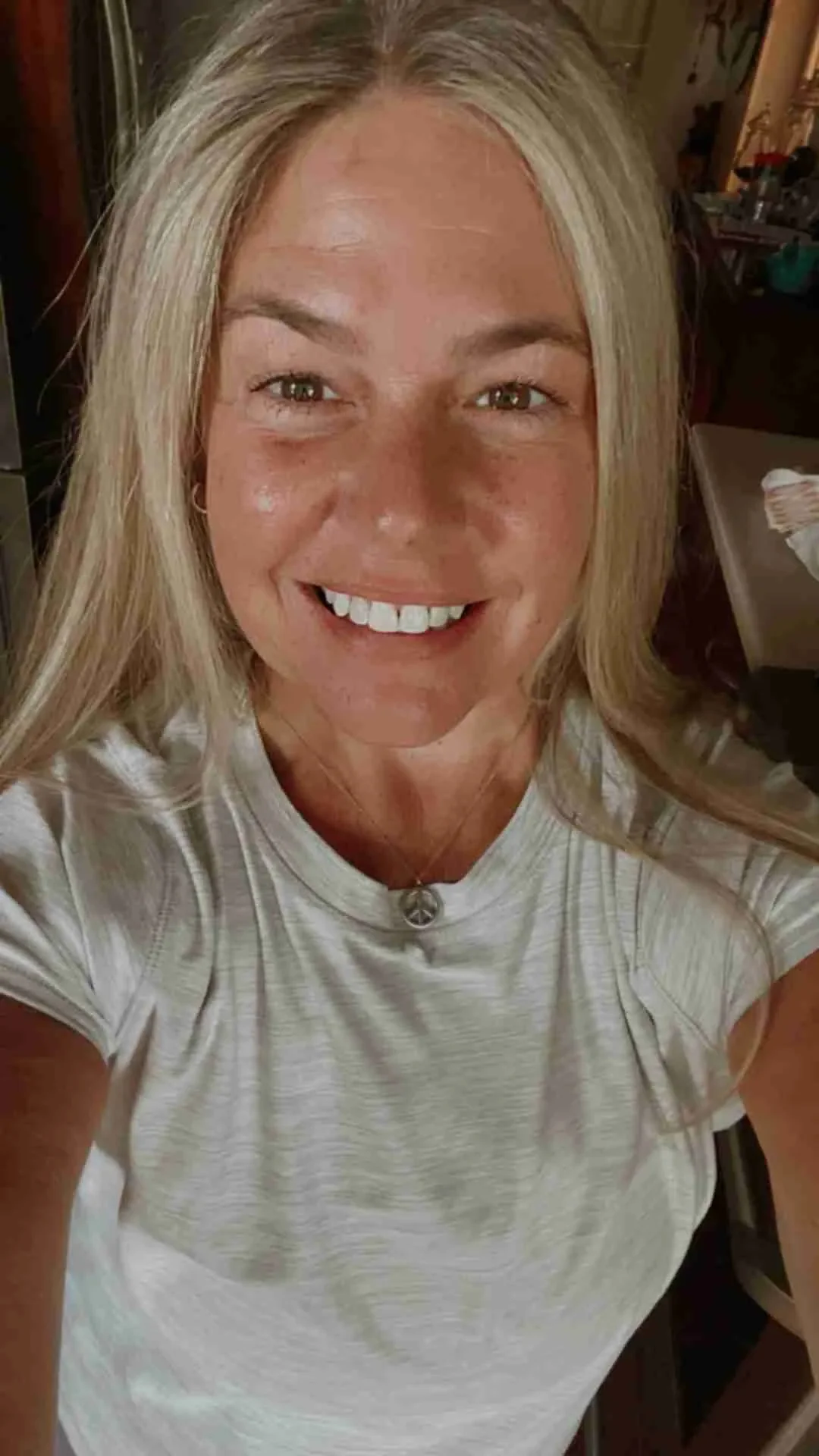 A woman with blonde hair smiling and taking a selfie, wearing a light-colored striped t-shirt and a peace sign necklace in a room with various objects in the background. CranialSacral Therapist at Spirit Well Sensory Lounge in Stuart, Florida.