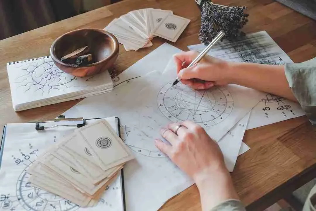 Person writing on astrology or astronomy charts with various papers, cards, and a small bowl on a wooden table.