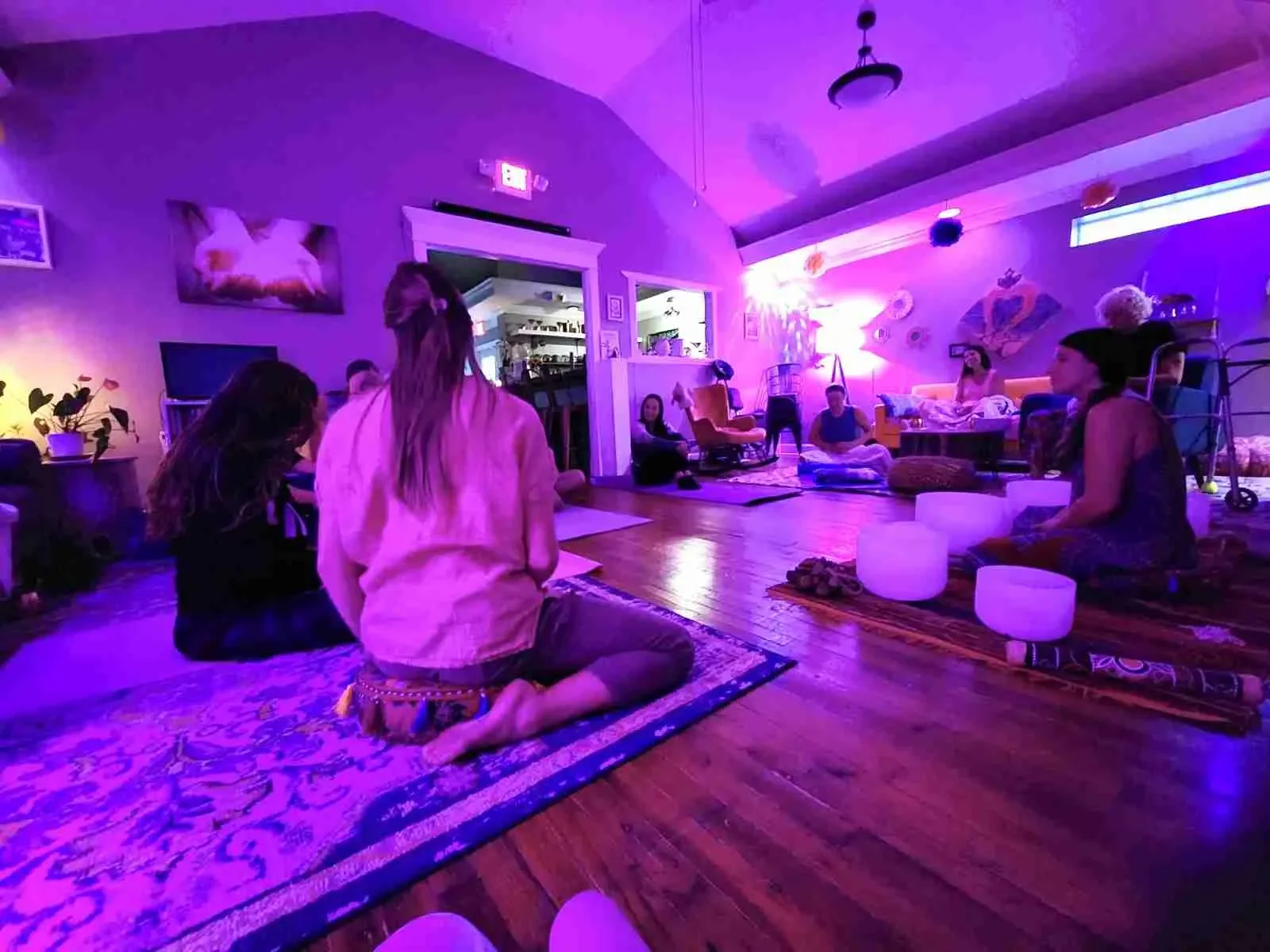 People participating in a meditation session in a dimly lit room with purple lighting, sitting on rugs on the wooden floor, some using singing bowls, with peaceful decorations on the walls. Sound Therapy at Spirit Well Sensory Lounge at Stuart, Fl.