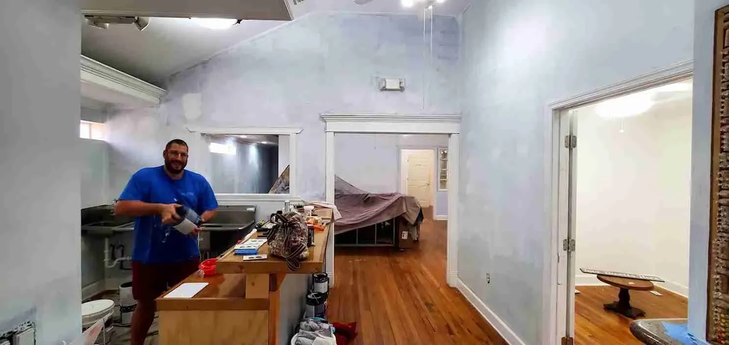 A man smiling and holding a drill tool in an unfinished or renovated room with light blue walls, hardwood floors, and open doorways. There is a counter with various items, and a bed covered with a brown sheet in the background.
