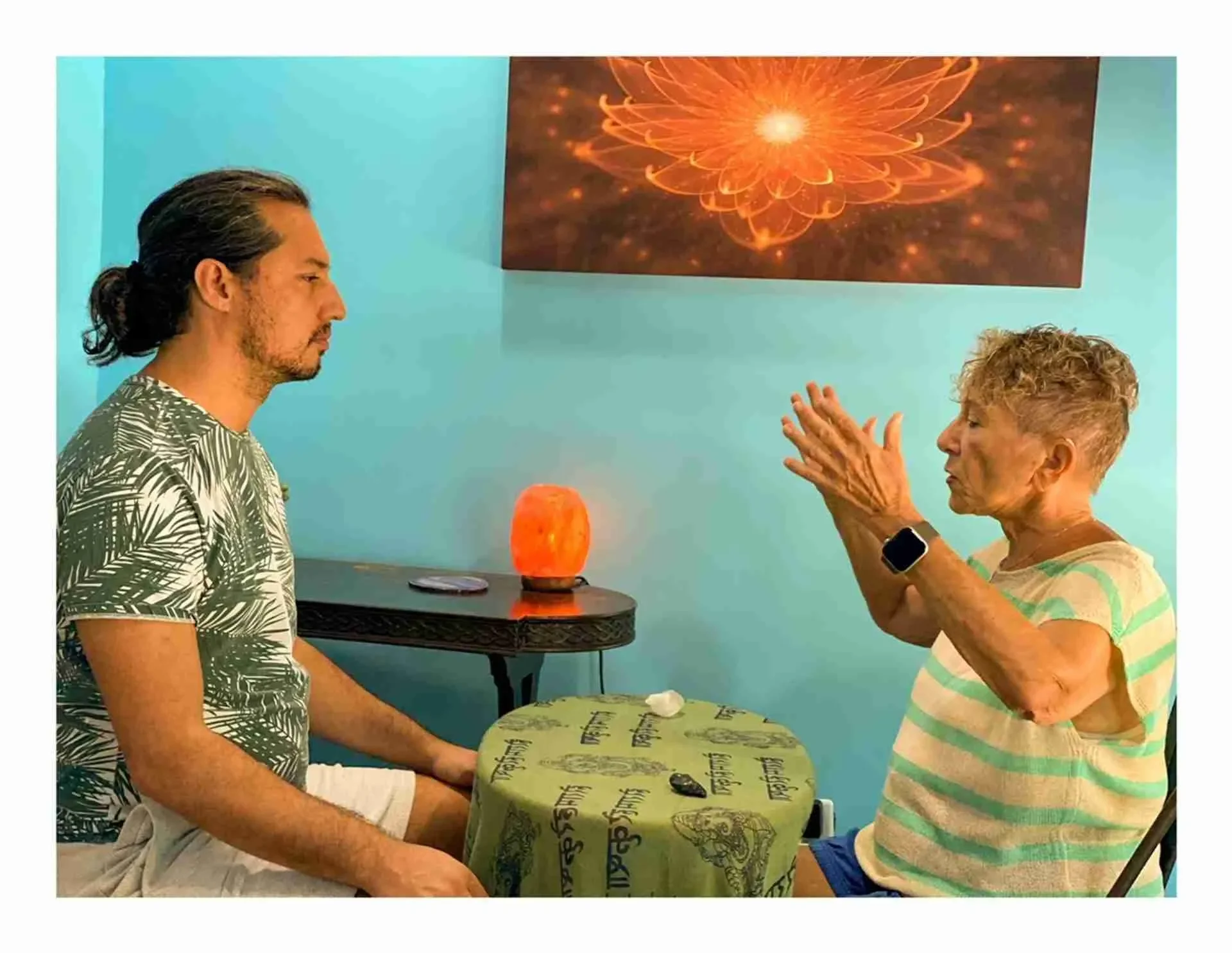 An elderly woman and a man sit across from each other at a small table with a green tablecloth in a room with turquoise walls. Marie Seger doing intuitive medicine counselling at Spirit Well Sensory Lounge in Stuart, Florida.