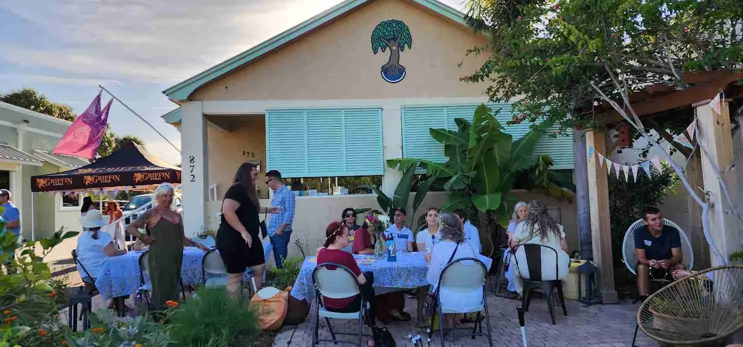 Group of people gathered around a table outdoors at an event, with some standing and some sitting, under a patio pergola with bunting decorations. There is a house with painted blue shutters and a tree with big leaves in the background.