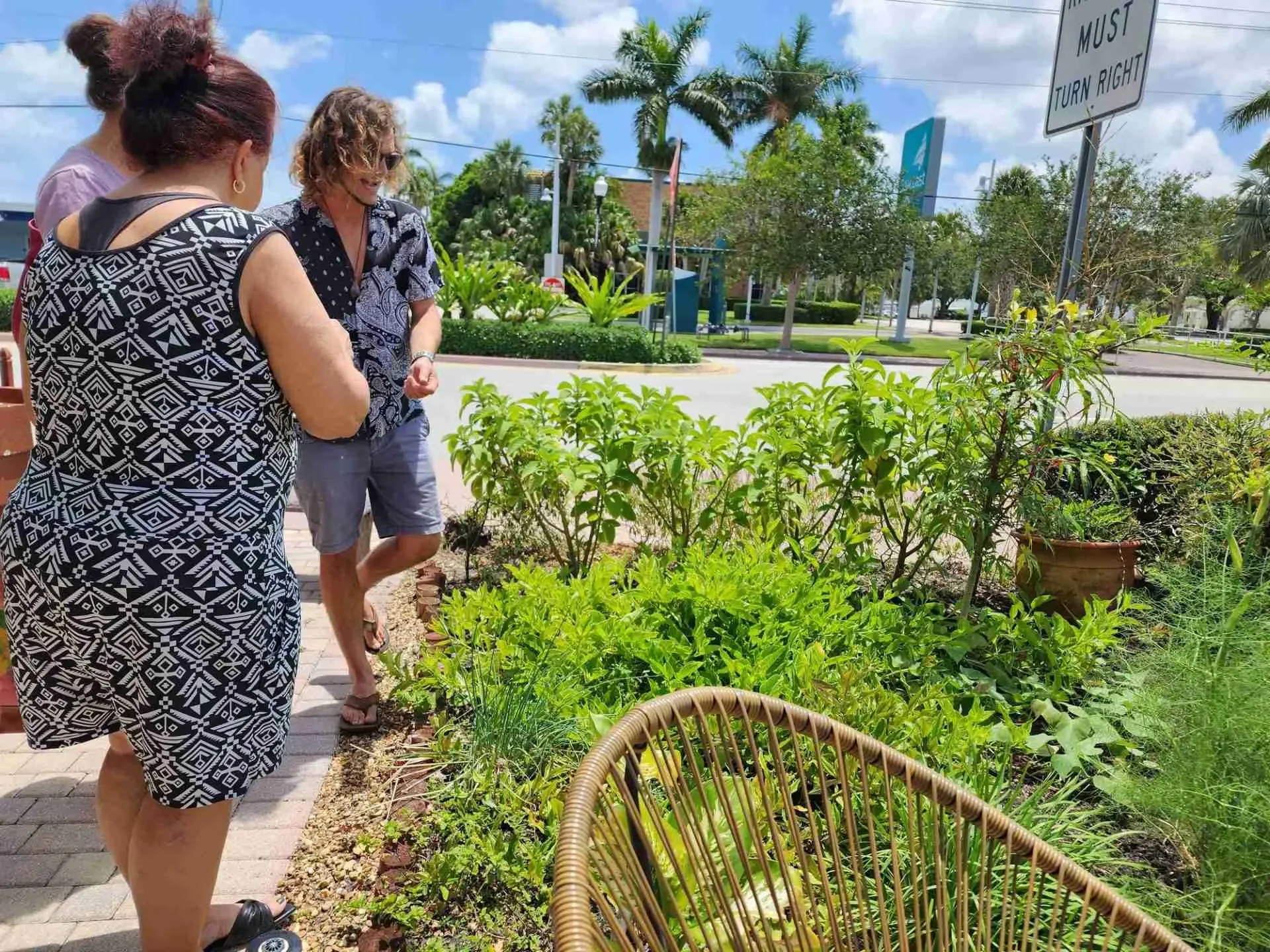 Three women looking at a garden of lush green plants outside on a sunny day, with palm trees, a street, and blue sky in the background. Outdoor area at Spirit Well Sensory Lounge in Stuart, Florida.