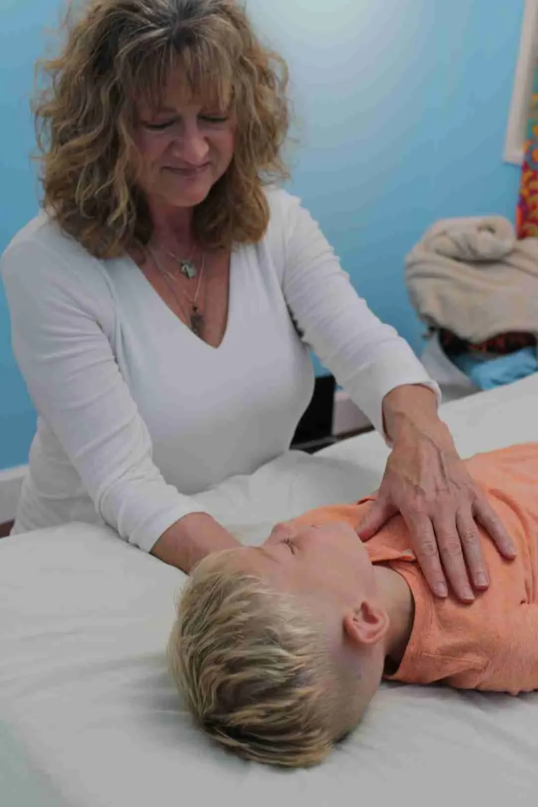 A woman giving a gentle chest compressions to a young boy lying on a hospital bed, both with light hair and the woman with curly hair, in a bright room with blue walls.