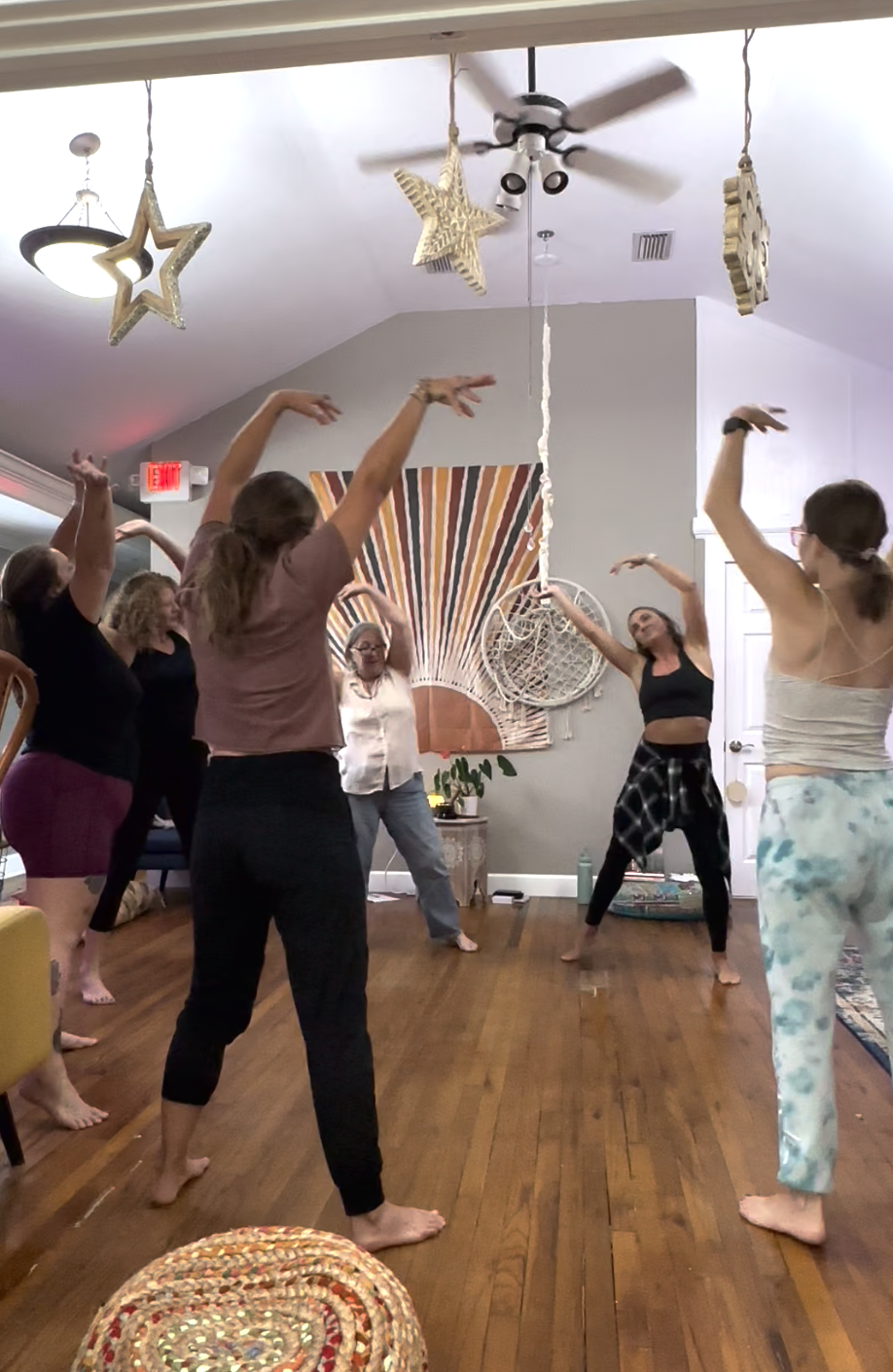 Group of women practicing yoga or stretching indoors, with decorative hanging stars and a large starburst art piece on the wall. Dance events and workshops at Spirit Well Sensory Lounge in Stuart, Florida.