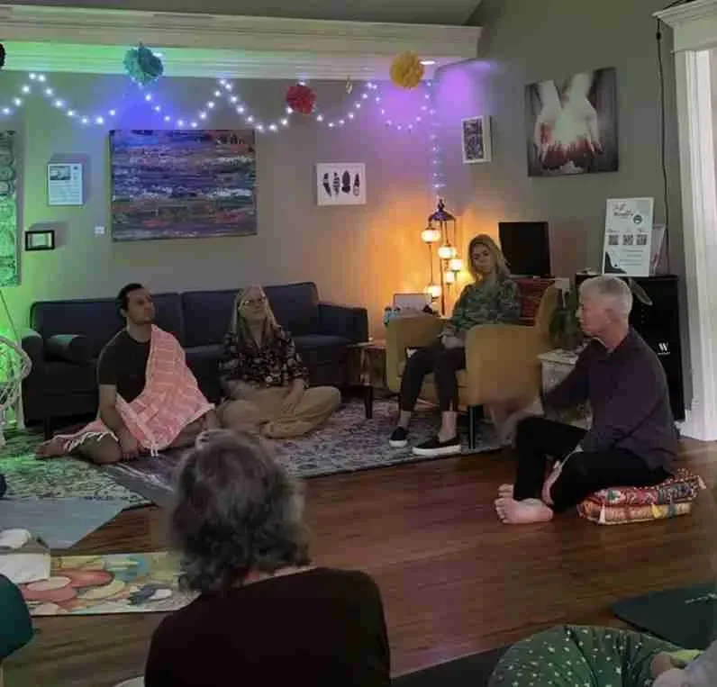 People sitting on the floor and in chairs in a decorated living room, with string lights and hanging paper decorations, participating in a mindfulness or meditation session. Spirit Well Sensory Lounge in Stuart, Florida.