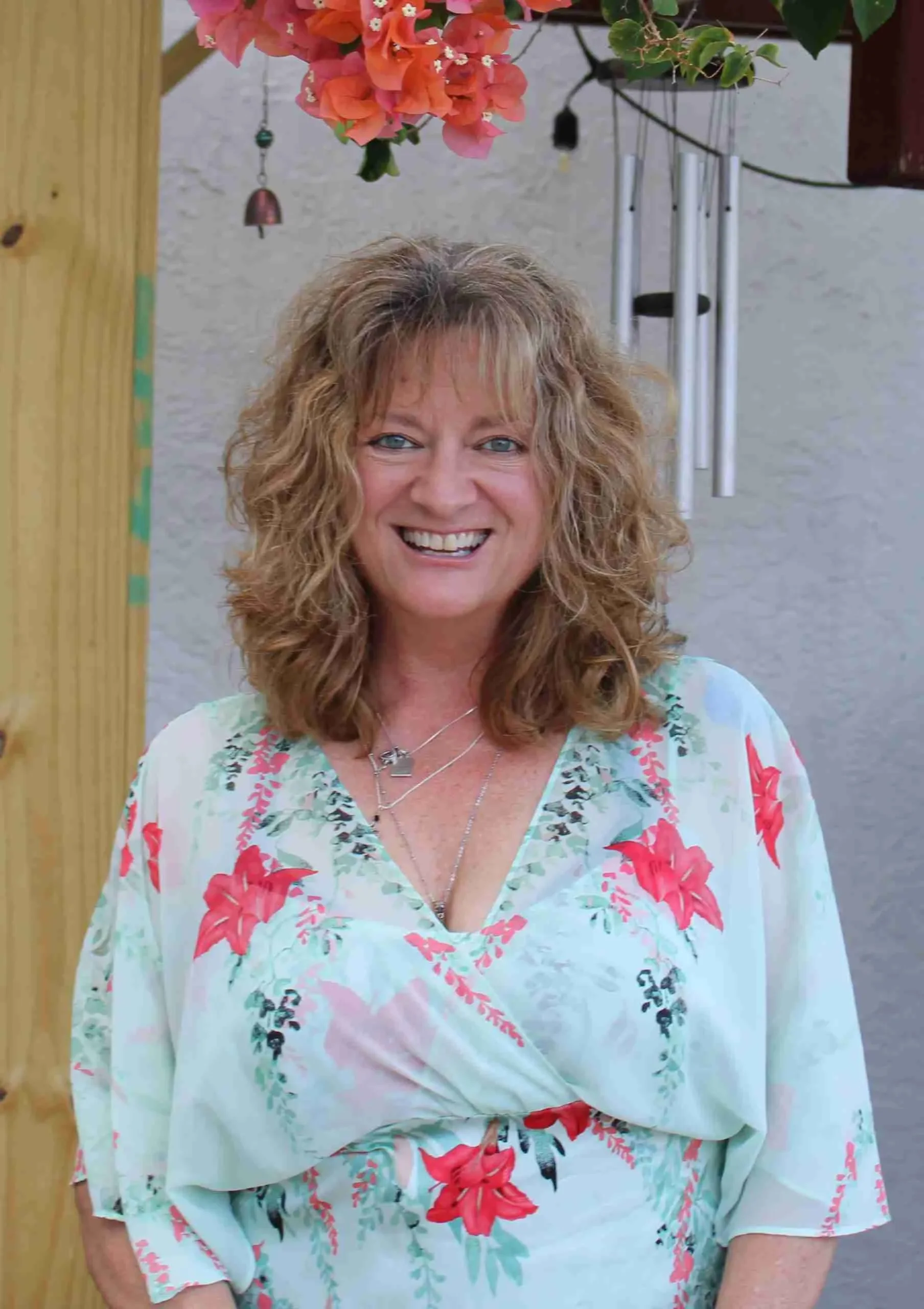 A woman with curly, shoulder-length blonde hair, wearing a floral blouse with red and pink flowers, smiling outdoors next to a wooden post and hanging wind chimes, Amy doing CranialSacral Therapy at Stuart, Florida in Spirit Well Sensory Lounge.