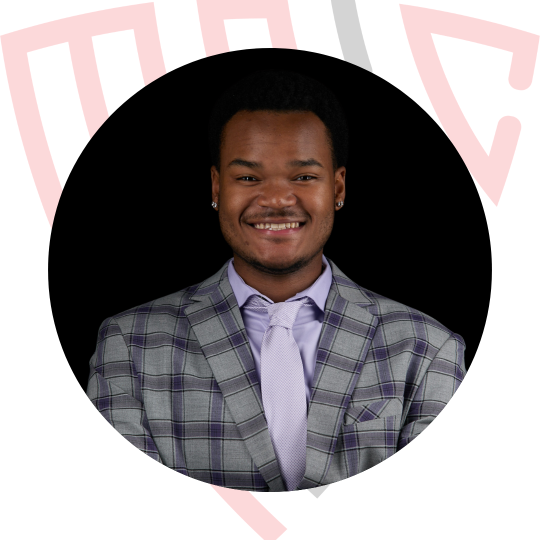 Young man in a gray suit and yellow tie, smiling, standing behind a clear podium with a microphone and papers, speaking at an event.