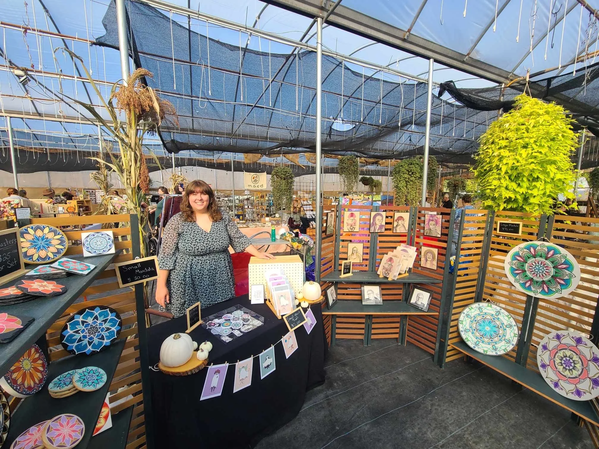 A woman standing behind a black display table with various handcrafted mandalas, art prints, and small pumpkins at a craft fair booth. The booth has decorative wooden panels and hanging plants, and other vendors are visible in the background under a large tent.