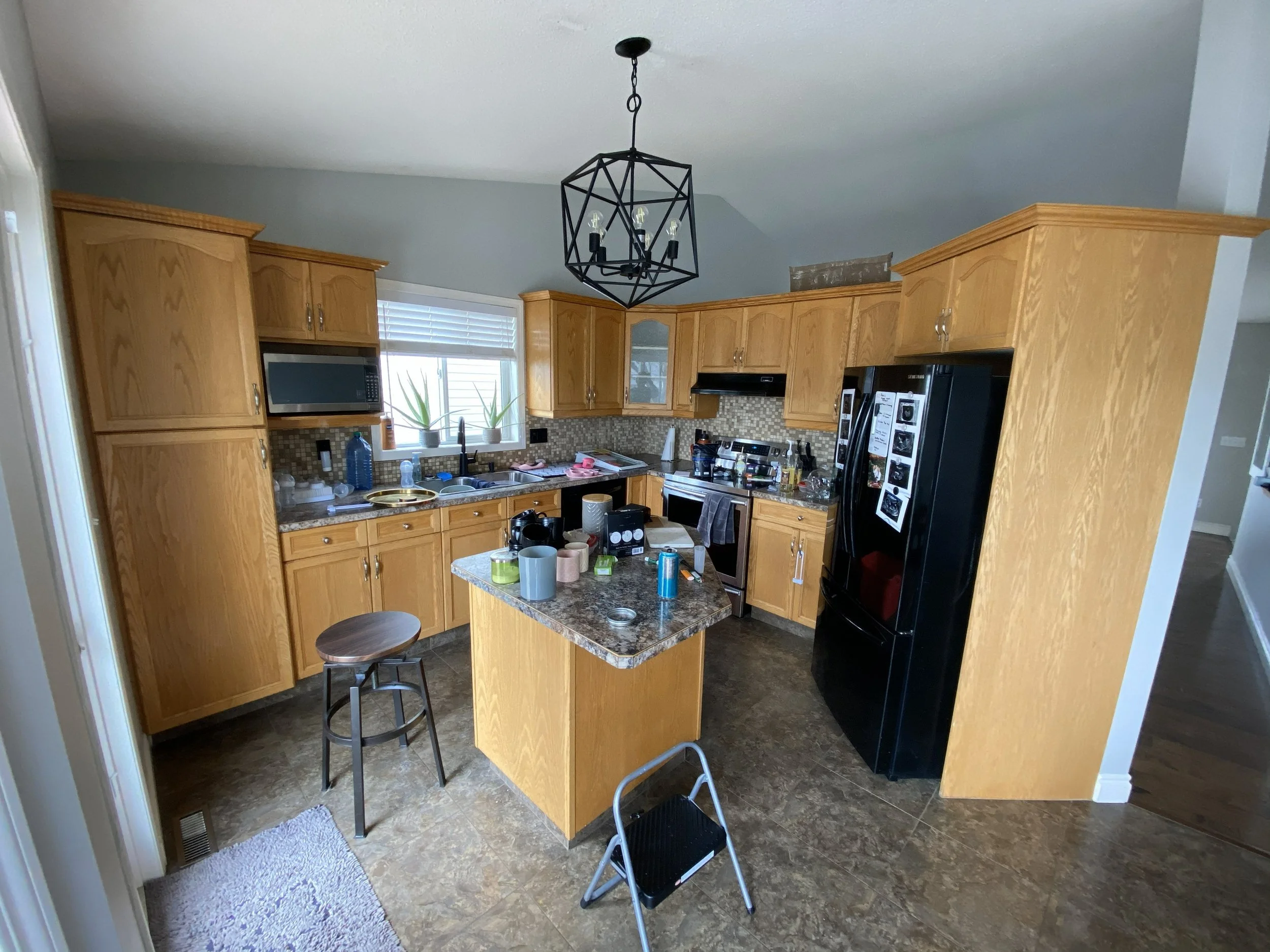 A cluttered kitchen with wooden cabinets, a black refrigerator, a mini island, and a window with blinds. The countertop has dishes, bottles, and various kitchen items.