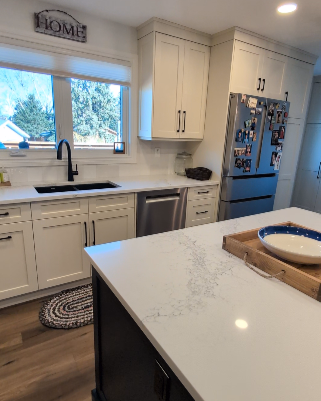 Kitchen with white cabinets, a window above the sink, stainless steel dishwasher, and a refrigerator covered with family photos. There is a white countertop with a blue and white bowl on a tray, and a small woven rug near the sink.