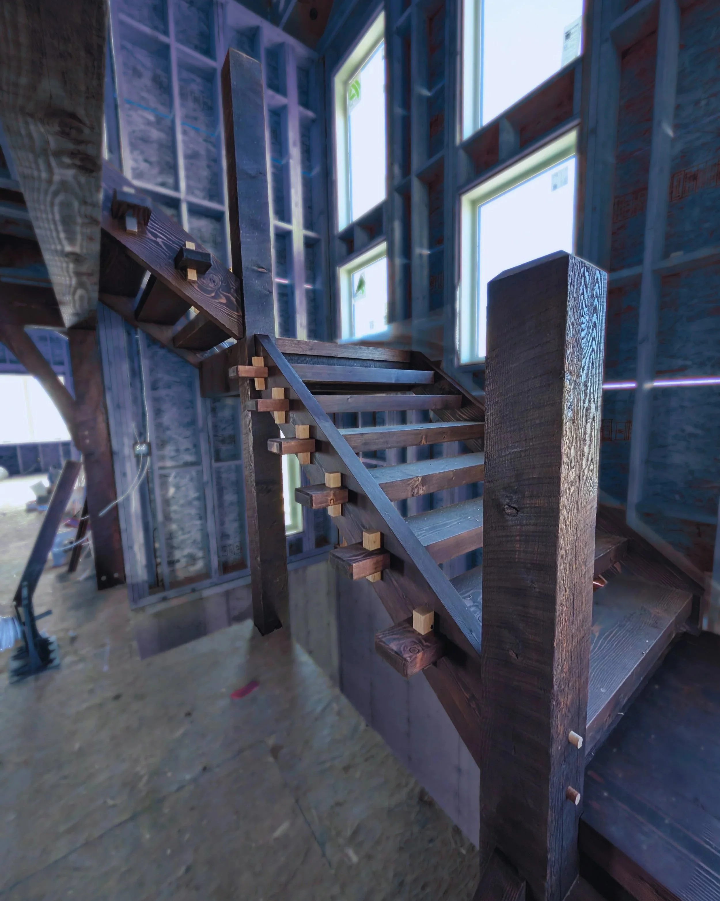 Interior of a house under construction with a wooden staircase and multiple window openings, showing exposed wall framing and insulation.