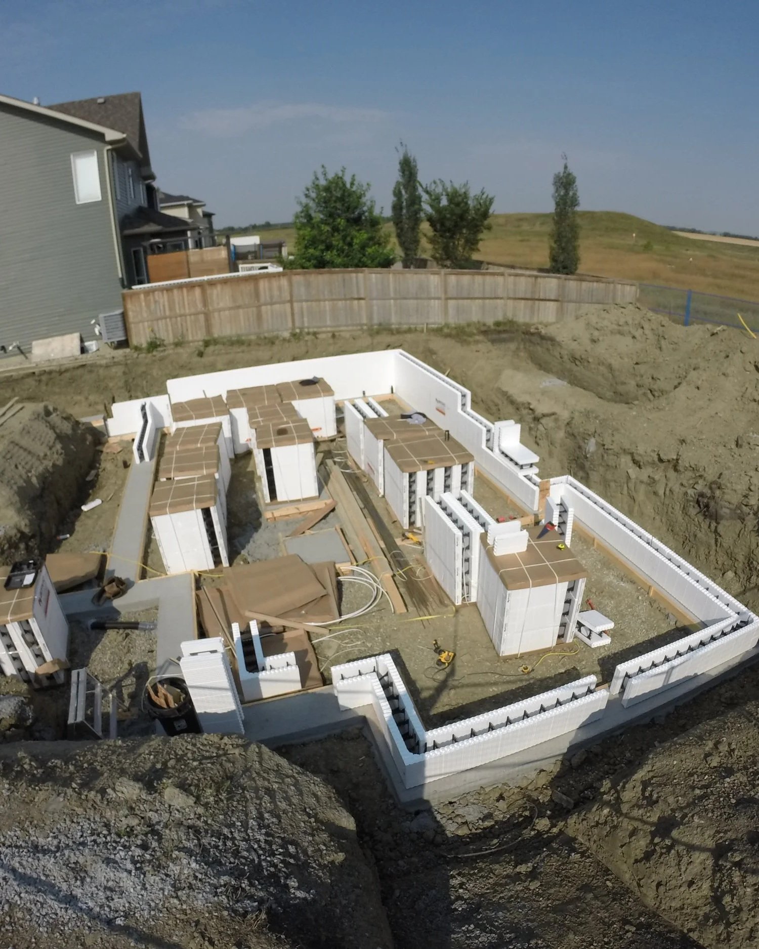 Foundation of a house under construction with white basement walls and plumbing, surrounded by dirt and construction equipment in an open yard.