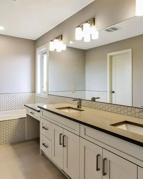 Modern bathroom with double vanity with two sinks, white cabinets, and a large mirror. There is a bathtub in the corner and a window providing natural light.