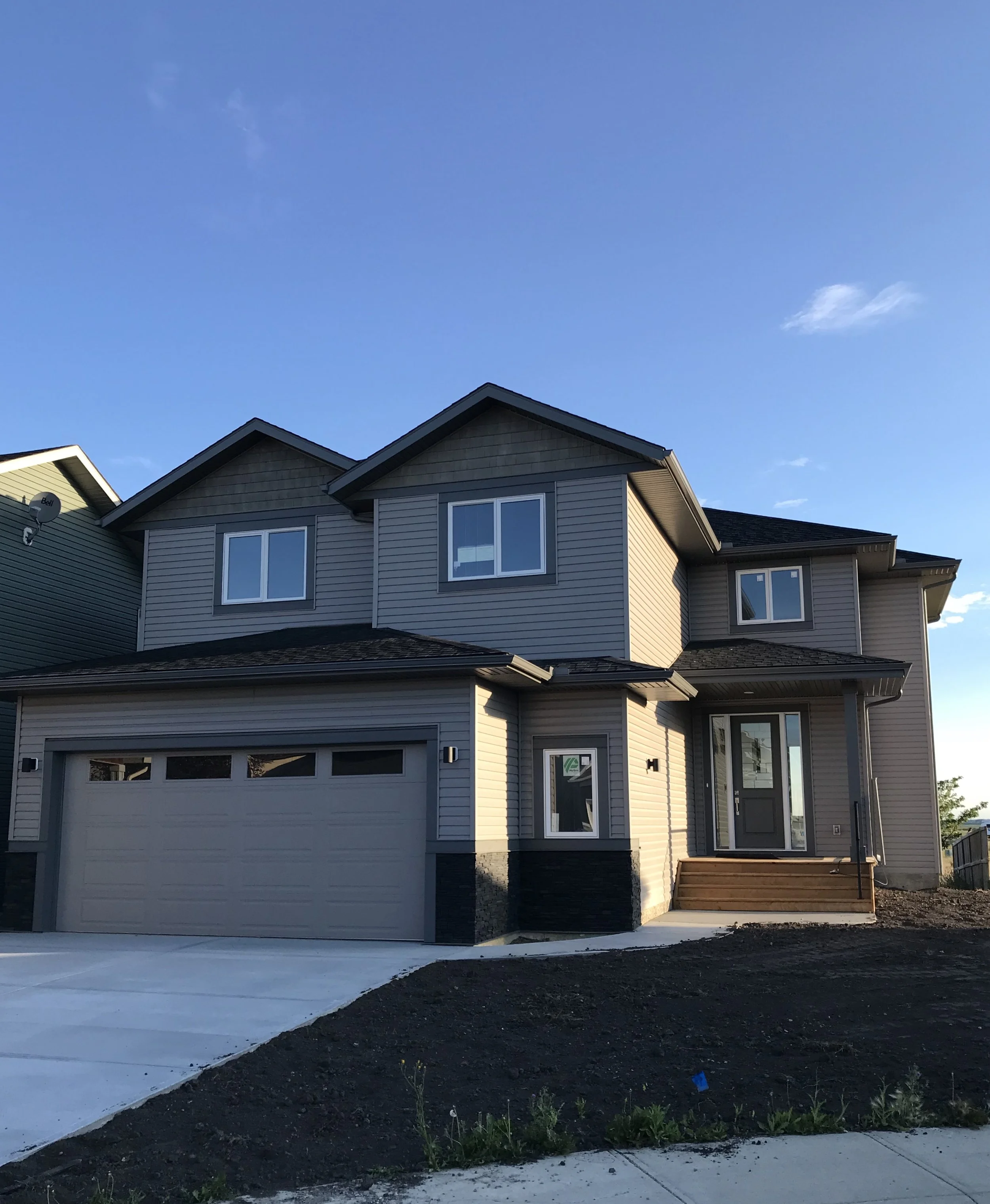 New two-story house with gray siding, black roof, main entrance door with steps, attached garage, and a small front yard with dirt and sparse grass, under a clear blue sky.